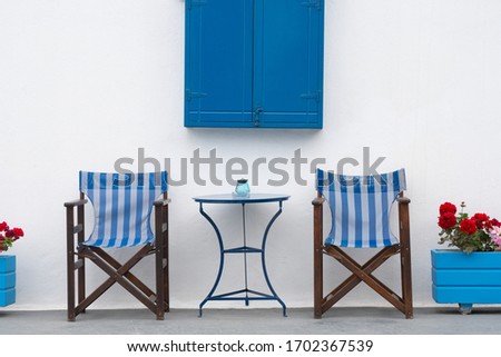 Relaxing balcony of greece taverna. Two blue chair facing the same direction in front of white cement wall and bright blue swing window