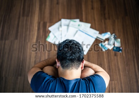 Top view of stressed young Asian man sitting and hugging knees, trying to find money to pay credit card debt and all loan bills. Financial problem from coronavirus or covid19 outbreak crisis concept. Royalty-Free Stock Photo #1702375588