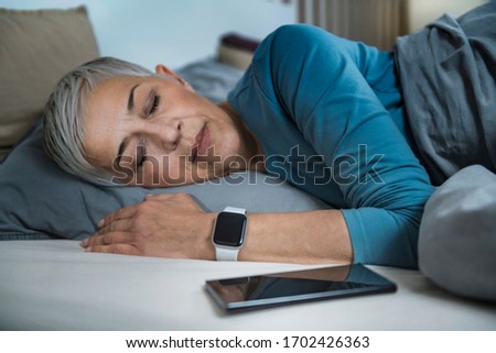 Sleep Apps - Tech-savvy Senior Woman Sleeping in Bed, Using Smart Phone and Smart Watch to Improve her Sleeping Habits Royalty-Free Stock Photo #1702426363