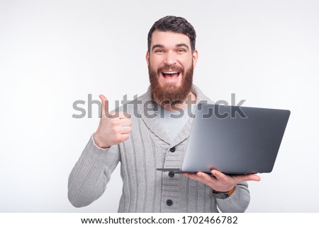amazed man with beard standing over white background while holding laptop and showing thumbs up