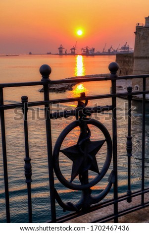 Railing with anchor and star during a warm sunset and in the background the merchant port of Taranto. Silhouette of port cranes. Puglia, Italy