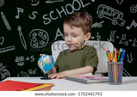 Cute clever boy is sitting at a desk with globe in hand on background with blackboard . Ready for school. Back to school.