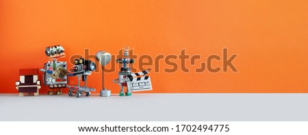 Robotic filmmaking backstage. Two robots shoots motion picture television episode or movie. Funny filmmakers director cameraman, cyborg assistant with clapperboard. Orange gray background, copy space  Royalty-Free Stock Photo #1702494775