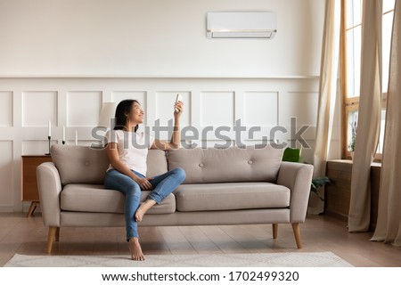 Young asian woman switching on air conditioner while resting seated on couch in modern interior living room, holds remote climate control cooler system manage temperature at contemporary home concept Royalty-Free Stock Photo #1702499320