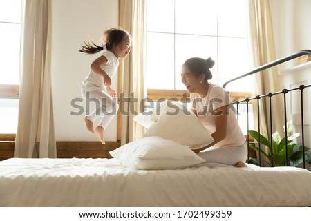 In morning carefree small daughter jump on bed while vietnamese mother laughing feels happy, asian ethnicity family in comfortable pyjamas wake up start new day positive mood enjoy active life concept Royalty-Free Stock Photo #1702499359