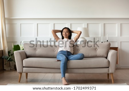 Asian woman looks at distance resting leaned on couch enjoy fresh air in summer day in modern fashionable living room interior, full length image. Contemporary apartments owner or carefree day concept Royalty-Free Stock Photo #1702499371