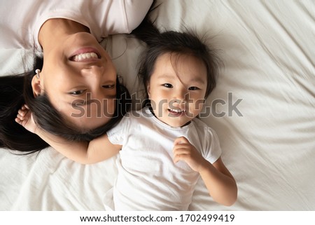 Asian single mother little daughter lying down together on bed white bedding smiling looking at camera feels joyful, above close up view. Happy motherhood, next generation, beautiful family portrait Royalty-Free Stock Photo #1702499419