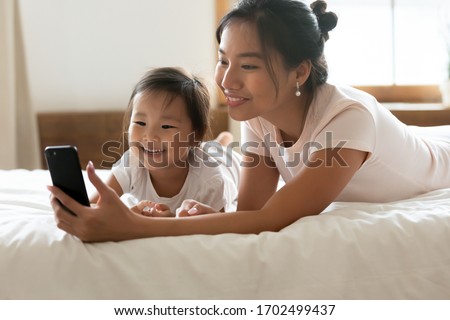Asian mum and little daughter in pyjamas after awakening have fun use cellphone watch cartoons, mom show kid girl educational on-line program. Parental control, modern tech, younger generation concept