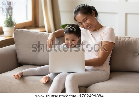 Parental control and educational website program children development concept. Asian mother her preschool little daughter wear comfy home clothes relaxing on couch with laptop having fun watch cartoon