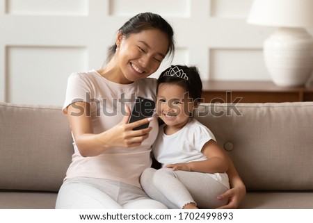 Cheerful Asian mother cuddles little sweet daughter holding smartphone making video call talk to relatives, taking selfie photography for memory, having fun at home use modern application tech concept