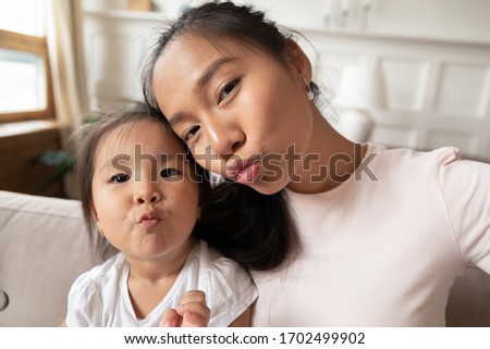 Beautiful asian mother and her little cute daughter taking selfie photography on gadget sending kisses making funny faces, webcam close up portrait view. Fun activity, modern tech young family concept