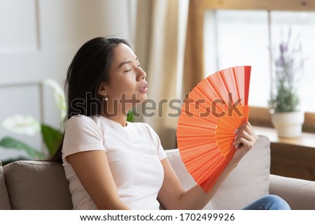 Overheated asian woman sweating feels discomfort seated on sofa at home without air-conditioning system. Tired girl suffers from heat stroke waving orange color fan cools herself in hot summer weather Royalty-Free Stock Photo #1702499926