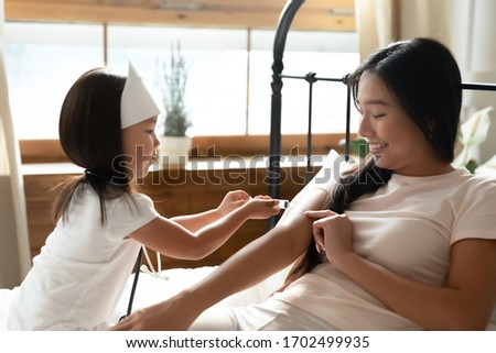 Asian little daughter plays doctor with mom seated on bed in bedroom, playful infant kid girl wearing nurse hat holding toy syringe gives injection to smiling young mother having fun together at home Royalty-Free Stock Photo #1702499935