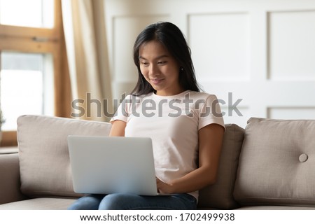 Asian young woman hold on lap notebook while typing e-mail seated on couch in living room alone. Social network media active user, do remote work, shopper makes goods services purchase on-line concept