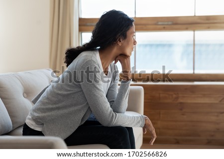Unhappy young african american girl sit on sofa at home and looking at window, thinking about problem and trouble, suffering from depression or loneliness, mental illness, makes difficult decisions Royalty-Free Stock Photo #1702566826