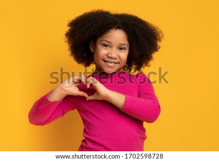 Cute little african american girl making heart shape with her hands and smiling to camera, posing over yellow background in studio, empty space Royalty-Free Stock Photo #1702598728