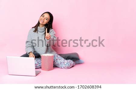 Young mixed race woman eating popcorn while watching a movie on the laptop showing and lifting a finger