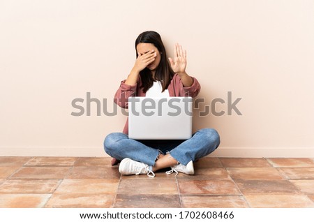 Young mixed race woman with a laptop sitting on the floor making stop gesture and covering face