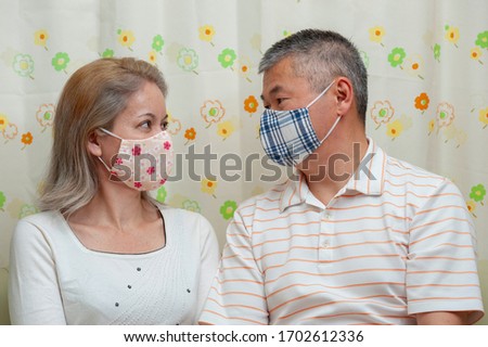 Middle-aged couple sitting looking at each other wearing homemade face mask (DIY) for protection against coronavirus (COVID-19). Floral curtain background. Royalty-Free Stock Photo #1702612336