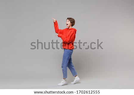 Side view of excited young brunette woman girl in casual red hoodie blue jeans posing isolated on grey background in studio. People lifestyle concept. Mock up copy space. Pointing index fingers aside
