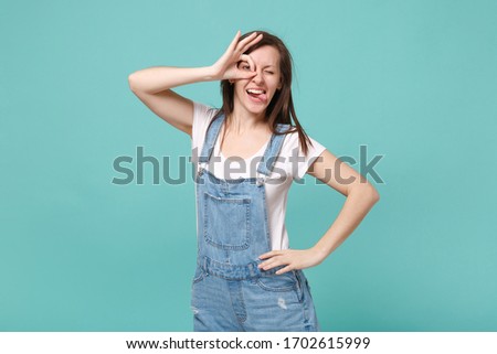 Funny young woman in casual denim clothes isolated on blue turquoise background. People lifestyle concept. Mock up copy space. Holding hands near eyes, imitating glasses or binoculars, showing tongue