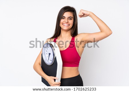 Young woman over isolated white background holding a weighing machine and doing strong gesture