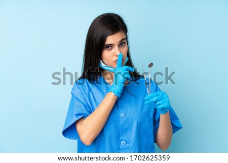 Young woman dentist holding tools over isolated blue background doing silence gesture