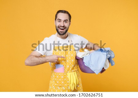 Smiling young man househusband in apron doing housework isolated on yellow background studio portrait. Housekeeping concept. Mock up copy space. Pointing index finger on basket with clean clothes