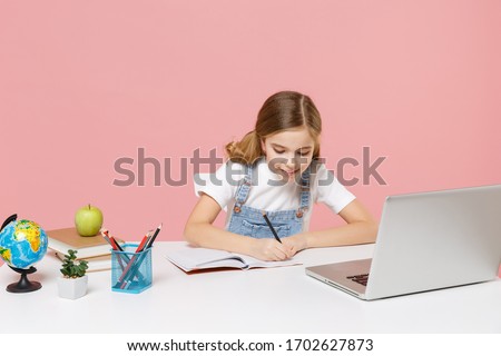 Smiling little kid schoolgirl 12-13 years old sit studying at white desk with pc laptop isolated on pink background. School distance education at home during quarantine concept. Writing in notebook