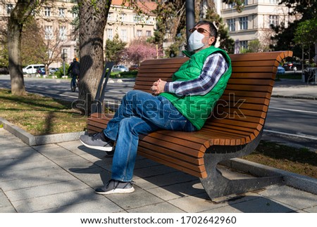 An elderly man in a medical mask sitting on a bench on a deserted embankment during a coronavirus epidemic in beautiful spring time