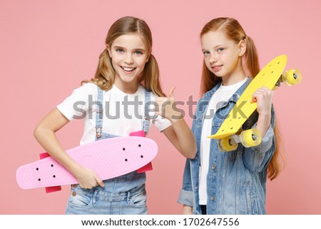 Smiling little kids girls 12-13 years old in white t-shirt, denim clothes isolated on pastel pink background studio. Childhood lifestyle concept. Mock up copy space. Hold skateboards showing thumb up