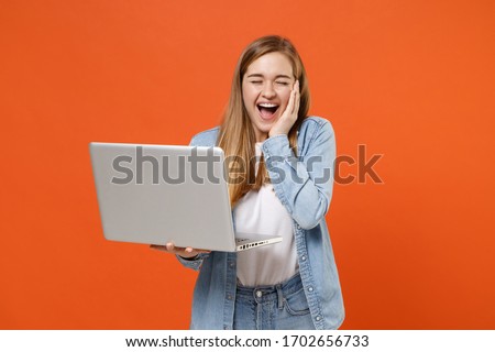 Funny young woman girl in casual denim clothes posing isolated on orange wall background. People lifestyle concept. Mock up copy space. Hold laptop pc computer, put hand on cheek, keeping eyes closed