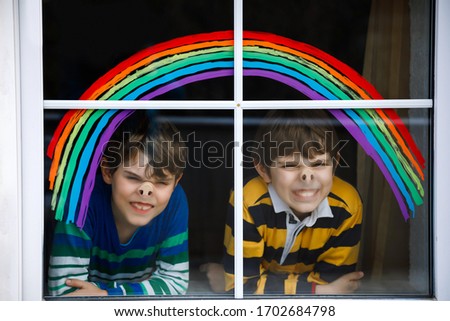 Two kids, school boys, siblings and best friends with rainbow painted with colorful window color during pandemic coronavirus quarantine. Children painting rainbows with the words Let's all be well