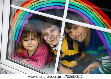 Three kids, two school kids boys and toddler girl with rainbow painted with colorful window color during pandemic coronavirus quarantine. Children painting rainbows with the words Let's all be well.