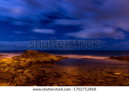 Long exposure shot of a cliff full of stones and a lagoon beside the ocean during a deep night as water poured onto the stones and the surrounding area.