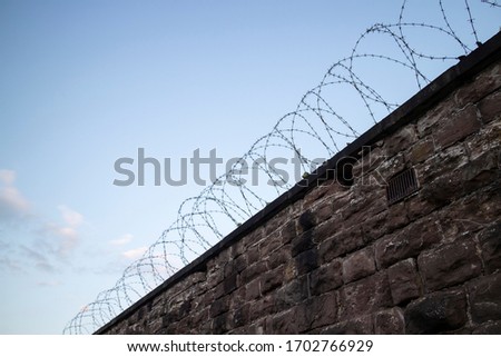 A grey stone wall, with a barbed wire fence on top, made to keep people out. The photo has a scary, sad tone to it. Although, there is a clear blue sky in the background. Angle is looking up at wall.