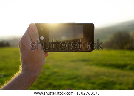 Taking photo with phone. A man's hand is holding a mobile smartphone and taking photo of the sun and sky in front of him. Man is white/Caucasian. Phone is black. Green field. It's a sunny, Summer day.