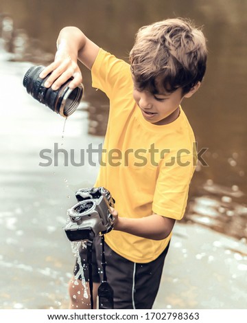 A schoolboy stands in the river on outdooor and destroys a mirrorless photo camera. The concept of obsolescence of technology. Photographers day, humor. Toned picture. Soft light photo.