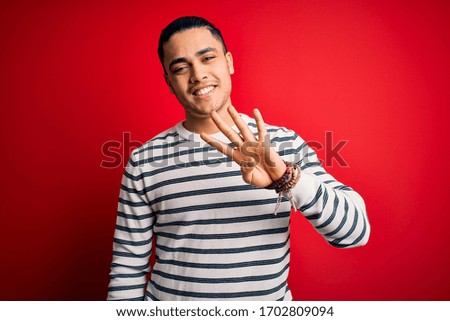 Young brazilian man wearing casual striped t-shirt standing over isolated red background showing and pointing up with fingers number four while smiling confident and happy.