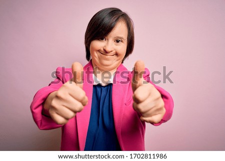 Young down syndrome business woman over pink background approving doing positive gesture with hand, thumbs up smiling and happy for success. Winner gesture.