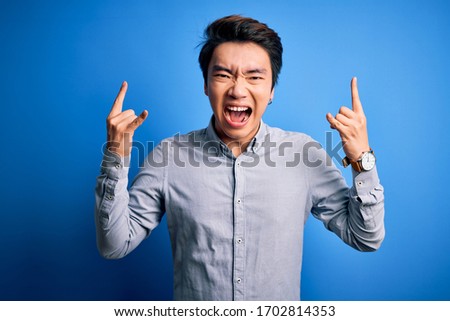 Young handsome chinese man wearing casual shirt standing over isolated blue background shouting with crazy expression doing rock symbol with hands up. Music star. Heavy music concept.