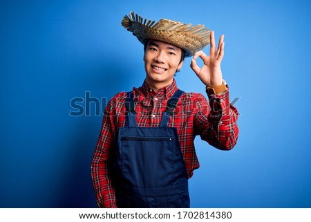 Young handsome chinese farmer man wearing apron and straw hat over blue background smiling positive doing ok sign with hand and fingers. Successful expression.