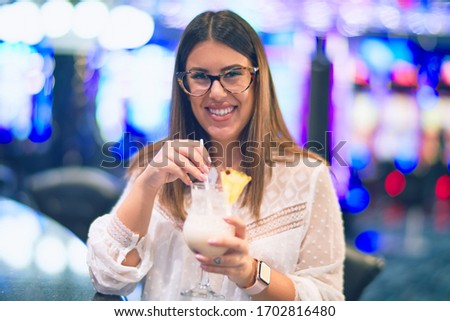 Young beautiful woman on vacation smiling happy and confident. Standing with smile on face drinking cocktail around lots of slot machine at casino