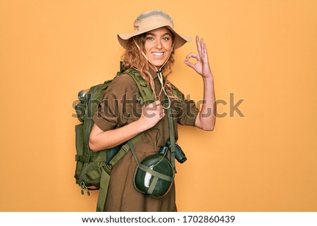 Young blonde explorer woman with blue eyes hiking wearing backpack and water canteen smiling positive doing ok sign with hand and fingers. Successful expression.