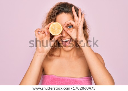 Beautiful woman with blue eyes wearing towel shower after bath holding lemon fruit over eye with happy face smiling doing ok sign with hand on eye looking through fingers
