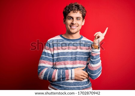 Young blond handsome man with curly hair wearing striped sweater over red background with a big smile on face, pointing with hand and finger to the side looking at the camera.