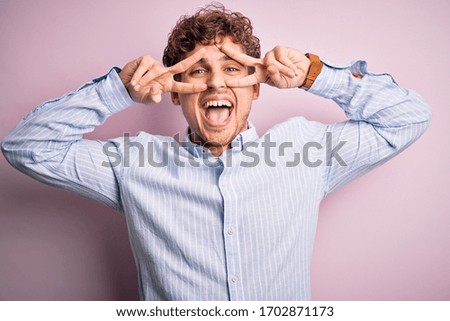 Young blond handsome man with curly hair wearing striped shirt over white background Doing peace symbol with fingers over face, smiling cheerful showing victory