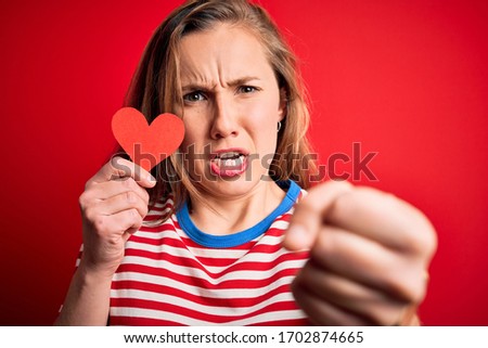 Young beautiful blonde woman holding paper heart standing over isolated red background annoyed and frustrated shouting with anger, crazy and yelling with raised hand, anger concept