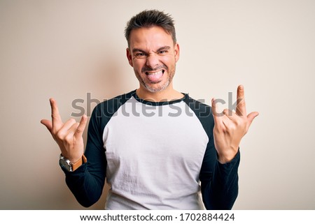 Young handsome man wearing casual t-shirt standing over isolated white background shouting with crazy expression doing rock symbol with hands up. Music star. Heavy concept.