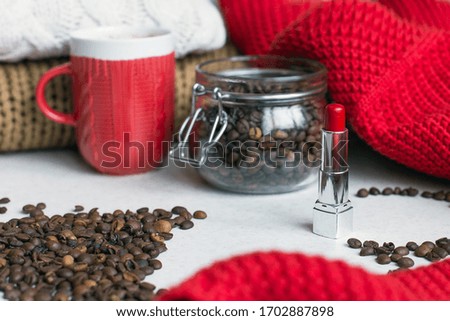 Macro shot of a transparent jar filled with coffee beans and a cup of coffee in the center of the photo, coffee beans around them and knitted sweaters in the background 